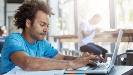 Stylish Afro American student keyboarding on laptop computer while sitting at cafe table with textbooks, working on homework, having focused concentrated look. People, modern technology and education